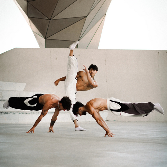 Three shirtless martial artists in white and black uniforms perform dynamic acrobatic karate poses in a large concrete courtyard under a geometric roof structure.