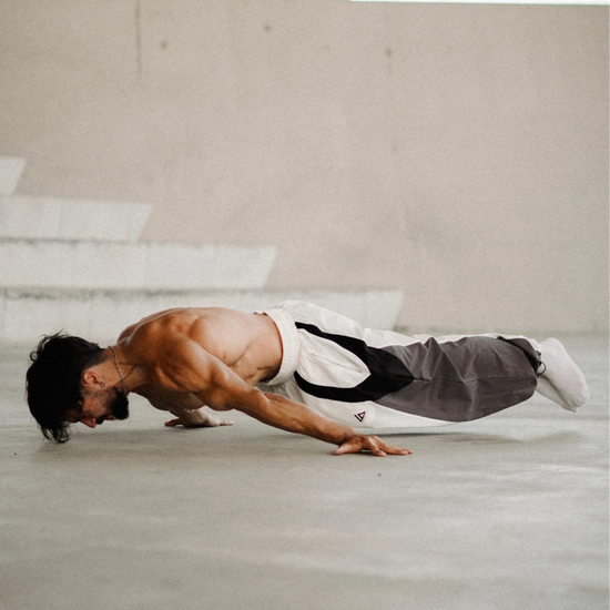 A shirtless man in a martial arts uniform performing a push-up on a concrete floor in front of a geometric modern building.
