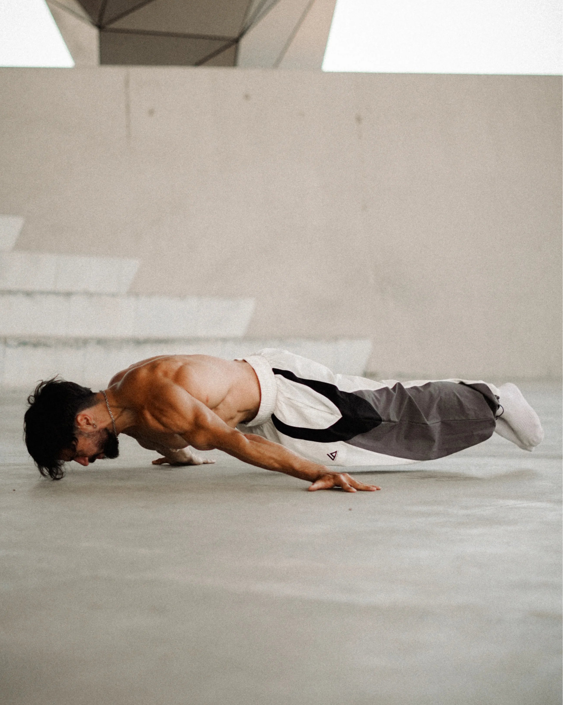 A shirtless man in a martial arts uniform performing a push-up on a concrete floor in front of a geometric modern building.