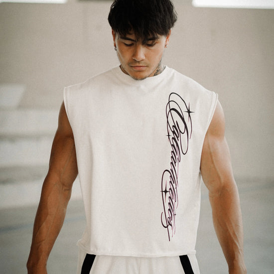 A muscular man wearing a sleeveless white athletic top with a black graphic print on the front, paired with white track pants with black side stripes, standing in a modern concrete structure.