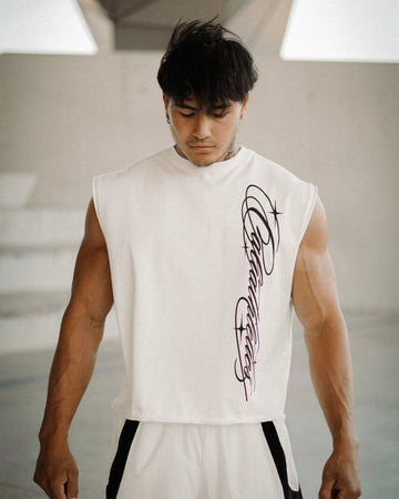 A muscular man wearing a sleeveless white athletic top with a black graphic print on the front, paired with white track pants with black side stripes, standing in a modern concrete structure.