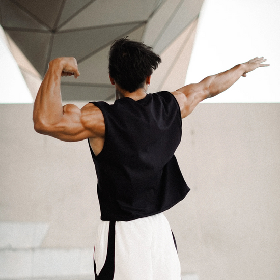 A muscular man shown from behind, flexing his bicep while wearing a black sleeveless shirt and white, gray, and black athletic pants in a modern concrete structure.