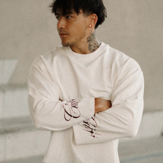 A man wearing a white long-sleeve crewneck sweater and black wide-leg pants stands with arms crossed in a modern concrete architectural setting.