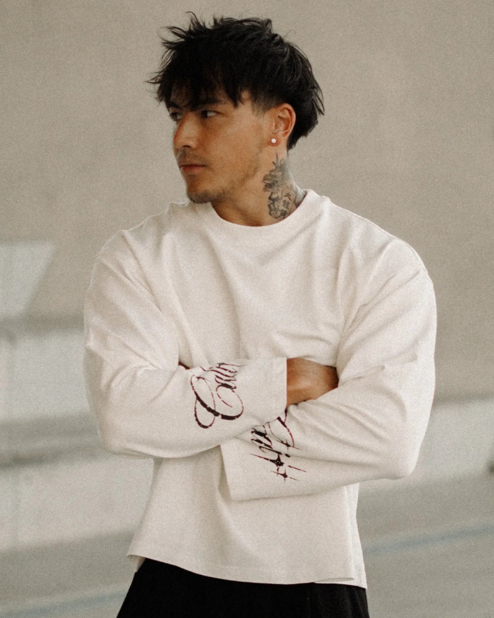 A man wearing a white long-sleeve crewneck sweater and black wide-leg pants stands with arms crossed in a modern concrete architectural setting.
