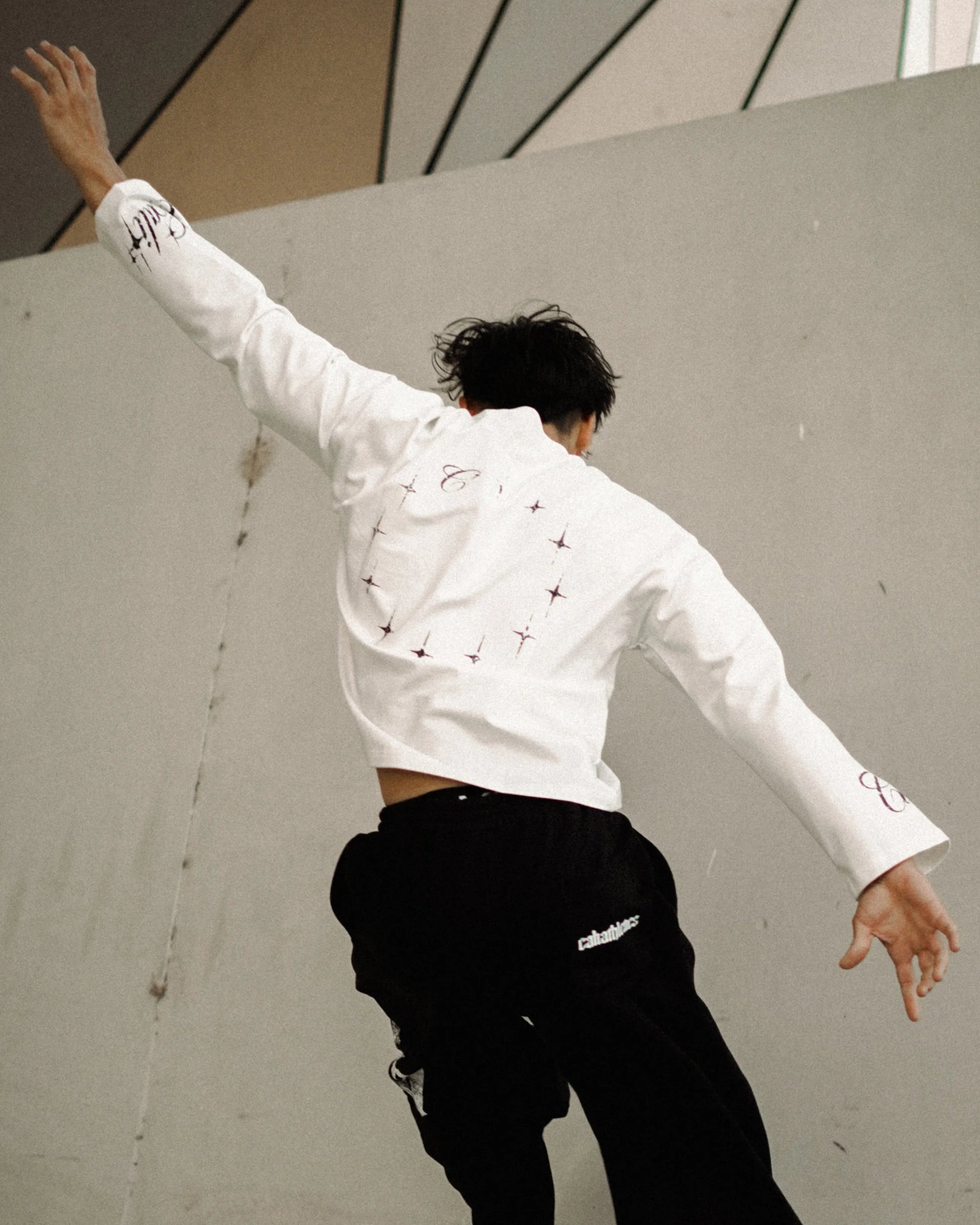A person wearing a white long-sleeve cropped shirt with small black details and black wide-leg pants is captured mid-motion against a large concrete wall in an outdoor urban setting.