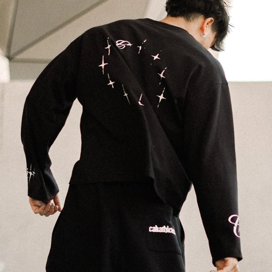 A man wearing a black streetwear outfit with white graphic details, captured from behind as he walks in front of a modern geometric concrete structure.
