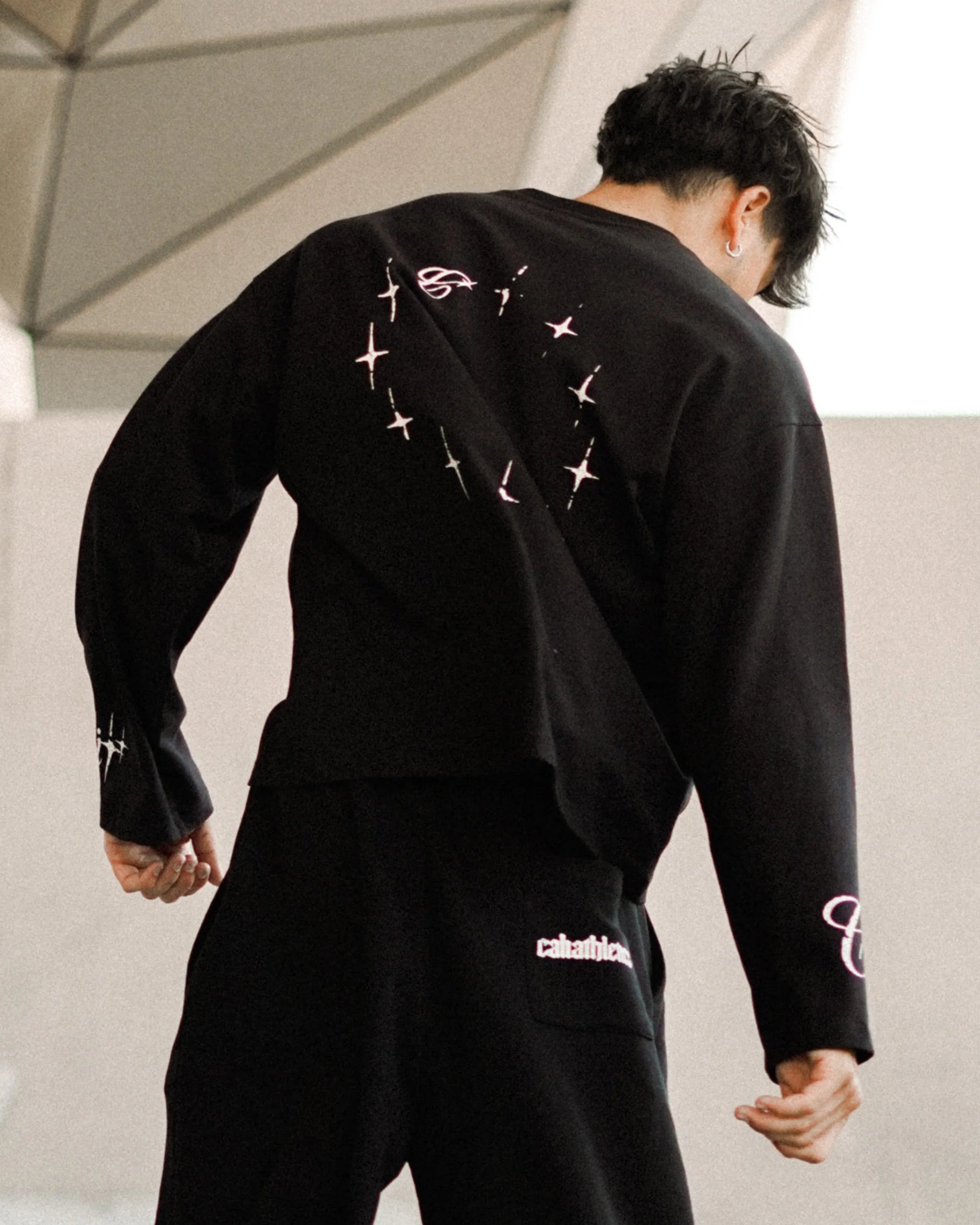 A man wearing a black streetwear outfit with white graphic details, captured from behind as he walks in front of a modern geometric concrete structure.